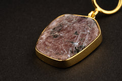 Close-up of raw Ruby pendant showing earthy red mineral texture, dark trace inclusions, gold-plated cap, and wide bail.