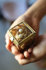 Small square premium gem display box held in hands, showing warm brass frame and transparent glass panels.