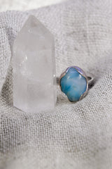 Oval Larimar ring shown on soft fabric beside a quartz crystal, with waved bezel and textured sterling silver band.
