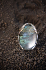 Oval Labradorite ring shown on earthy background, with waved bezel setting and shifting blue-green flash.