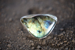 Triangular Labradorite ring shown on earthy background, with waved bezel setting and shifting gold-blue flash.