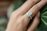 Long square cut Aquamarine sterling silver ring worn on hand against green leaves, showing hammered band and pale blue gemstone.