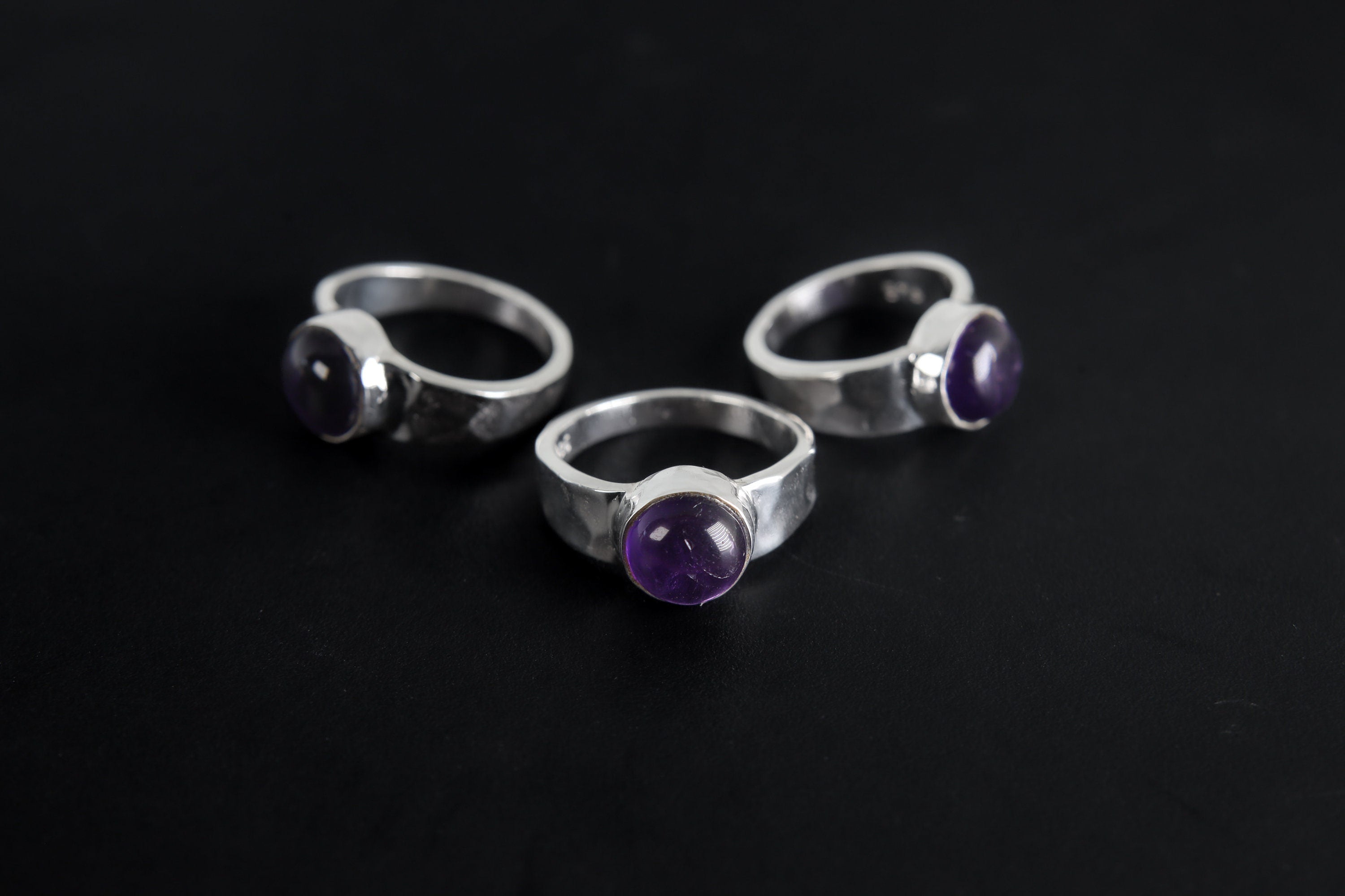 Group of Harmonious Amethyst Essence Rings shown on black background with hammered sterling silver bands and purple stones.