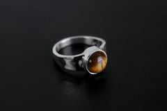 Amber Gaze Tiger Eye Ring shown on black background with round golden-brown cabochon and hammered silver band.