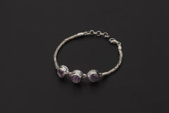 Amethyst bracelet displayed on dark background with three faceted purple stones and sterling silver Byzantine chain.