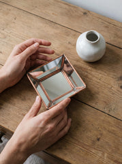 Handmade copper mirror styled on a wooden table, showing reflective square design and warm metal frame.