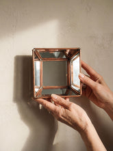 Handmade square copper mirror held in hand, showing angled geometric mirror panels and warm copper frame.