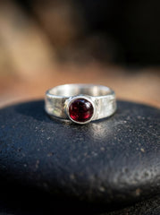Crimson Sentinel Garnet Ring shown on dark surface with polished red Garnet and hammered sterling silver setting.