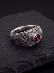 Garnet oxidized sterling silver ring displayed on dark slate with textured signet band and red gemstone.