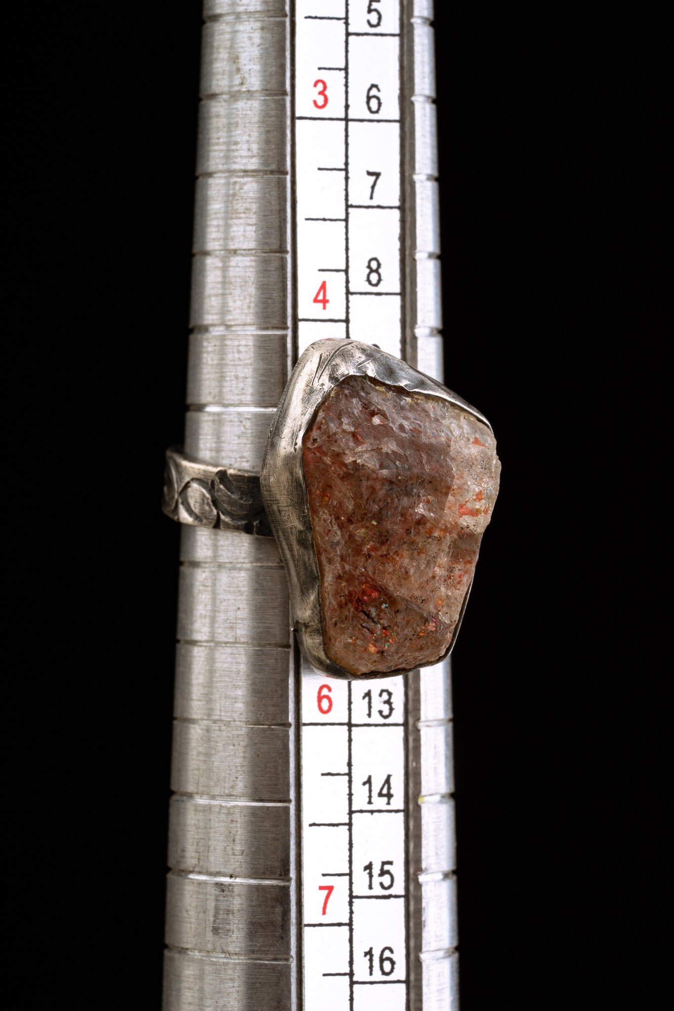 A silver ring with a large, irregularly shaped, clear, brown, and red gemstone. The ring is sized on a ring sizer that shows a size 4.5. to 5