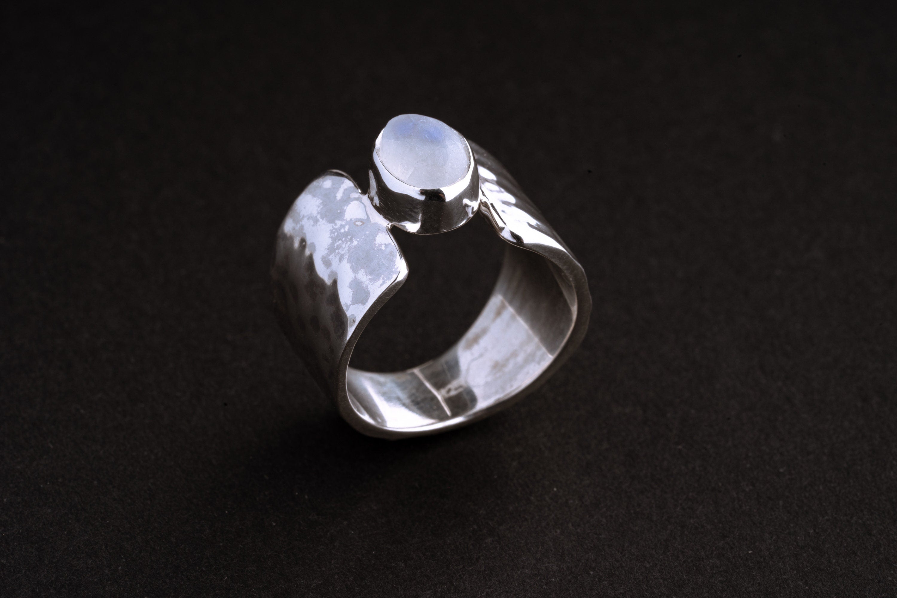 Blue Moonstone Hammered Silver Ring shown on black background with organic wide silver band and icy Moonstone centre.