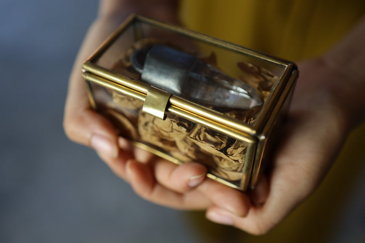 Large rectangular brass and glass display box held in hands with jewelry and crystal keepsakes inside.
