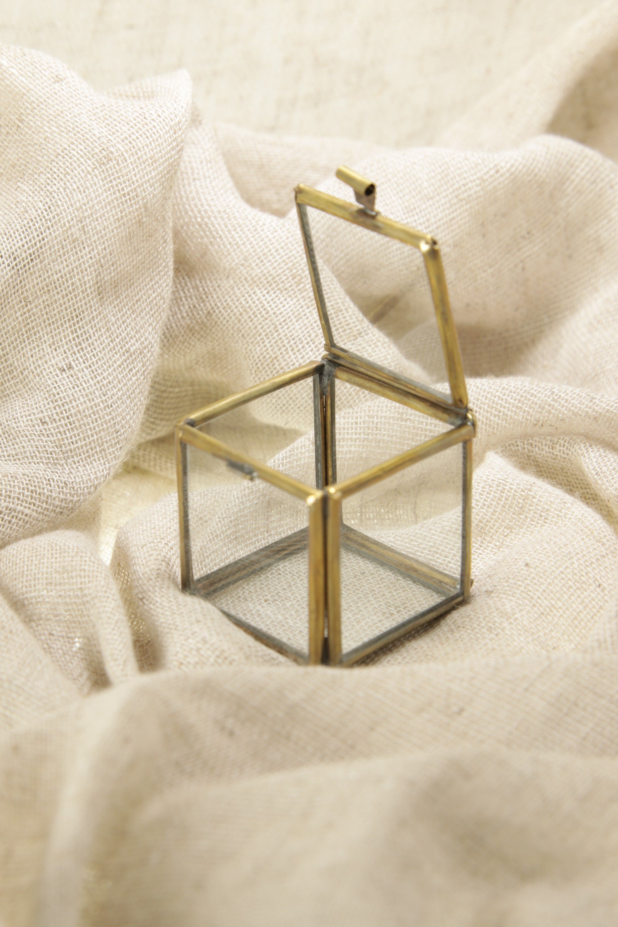 Gold-toned brass glass display box shown on soft fabric with open lid and clear square structure.