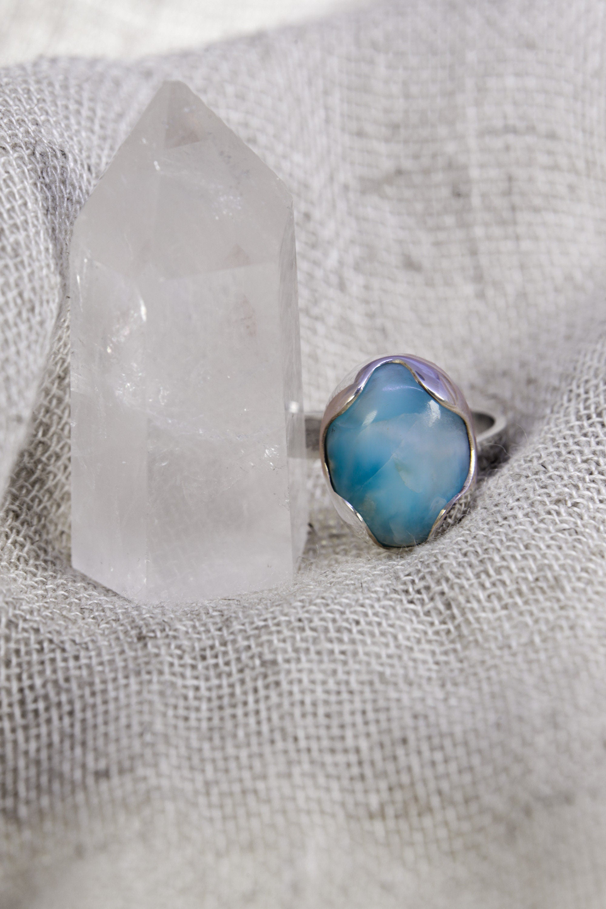 Oval Larimar ring shown on soft fabric beside a quartz crystal, with waved bezel and textured sterling silver band.