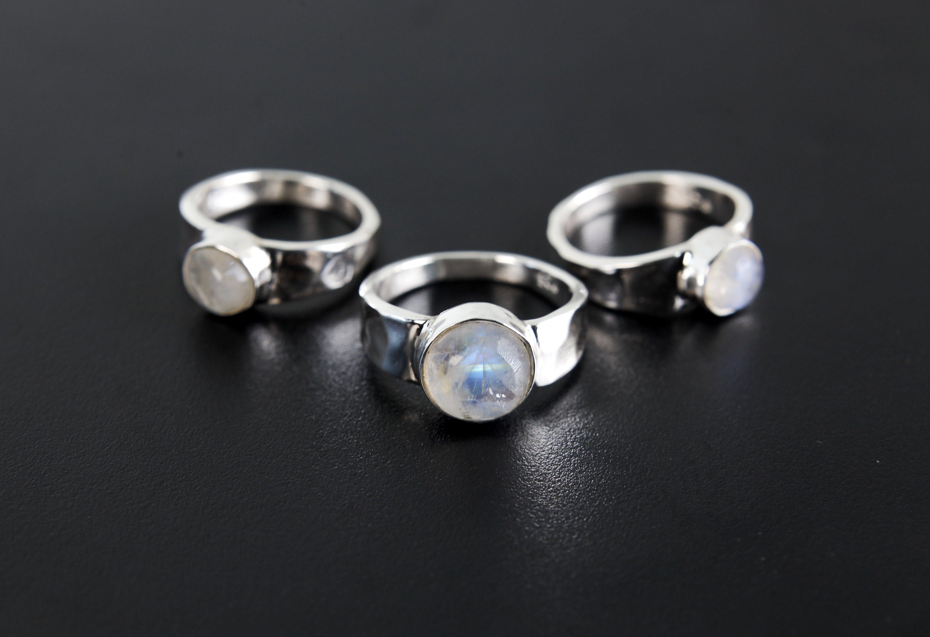 Group of Radiant Blue Moonstone Hammered Rings shown on black background with shiny sterling silver bands and round stones.