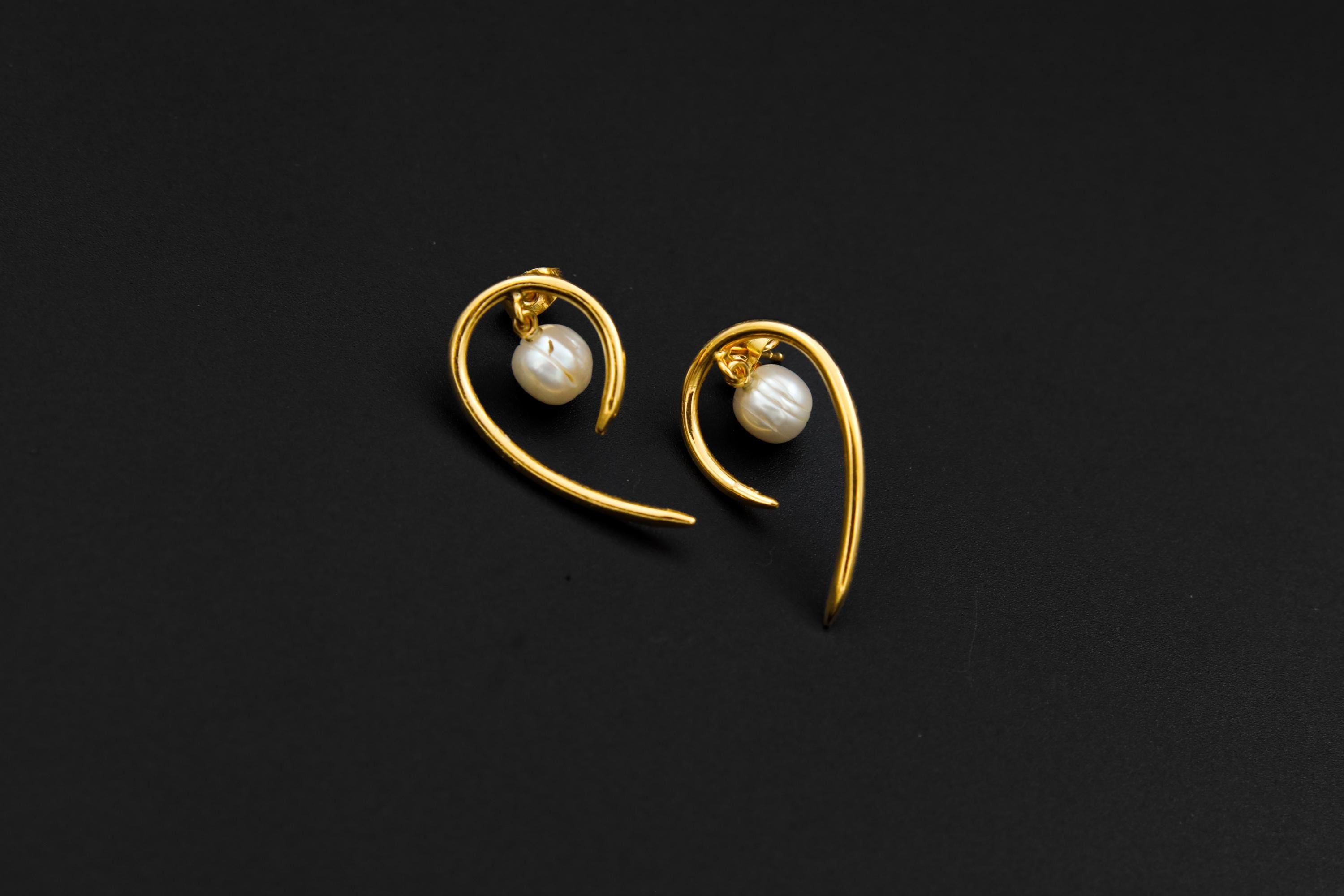 Gold-plated Pearl stud earrings shown on a dark background, with sculptural curled horn design and soft Pearl glow.