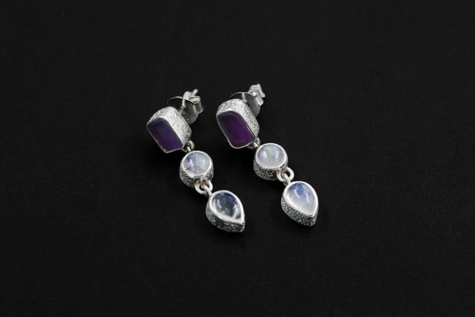 Handmade Amethyst and Moonstone sterling silver drop earrings shown on dark background with sand-textured bezels.
