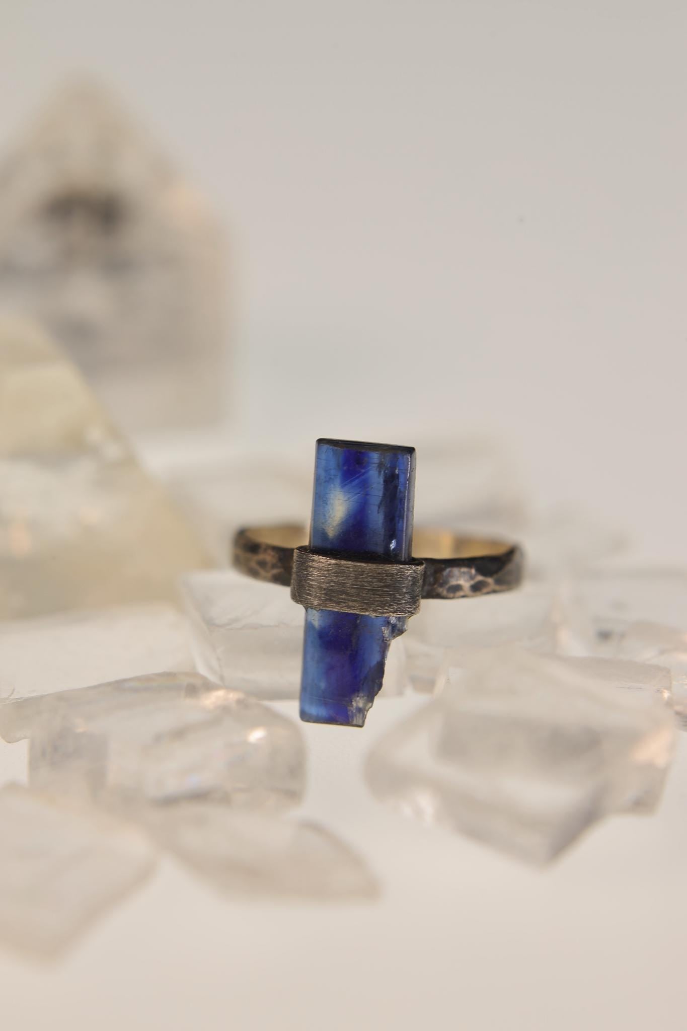 Raw Kyanite sterling silver ring displayed on clear quartz, with deep blue blade crystal and oxidized hammered band.