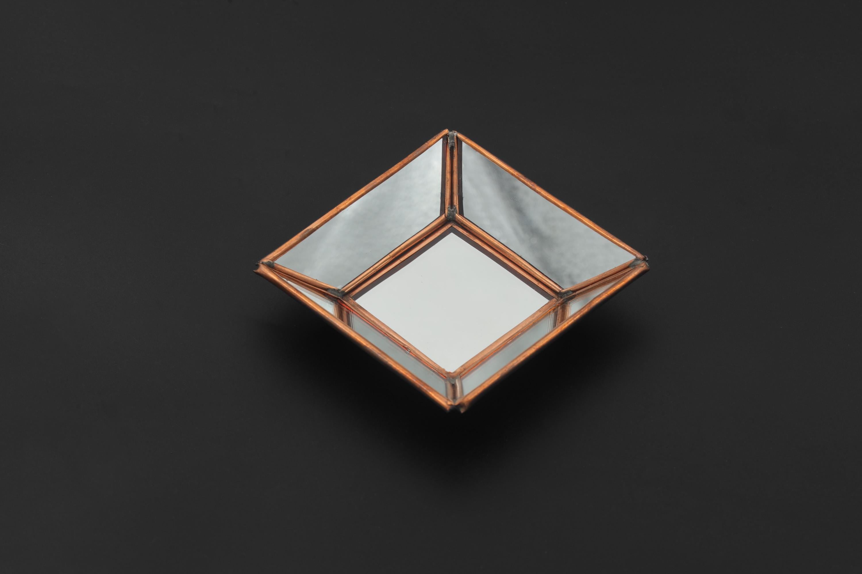 Square copper-framed mirror shown on dark background with angled mirrored glass panels and sculptural geometric form.