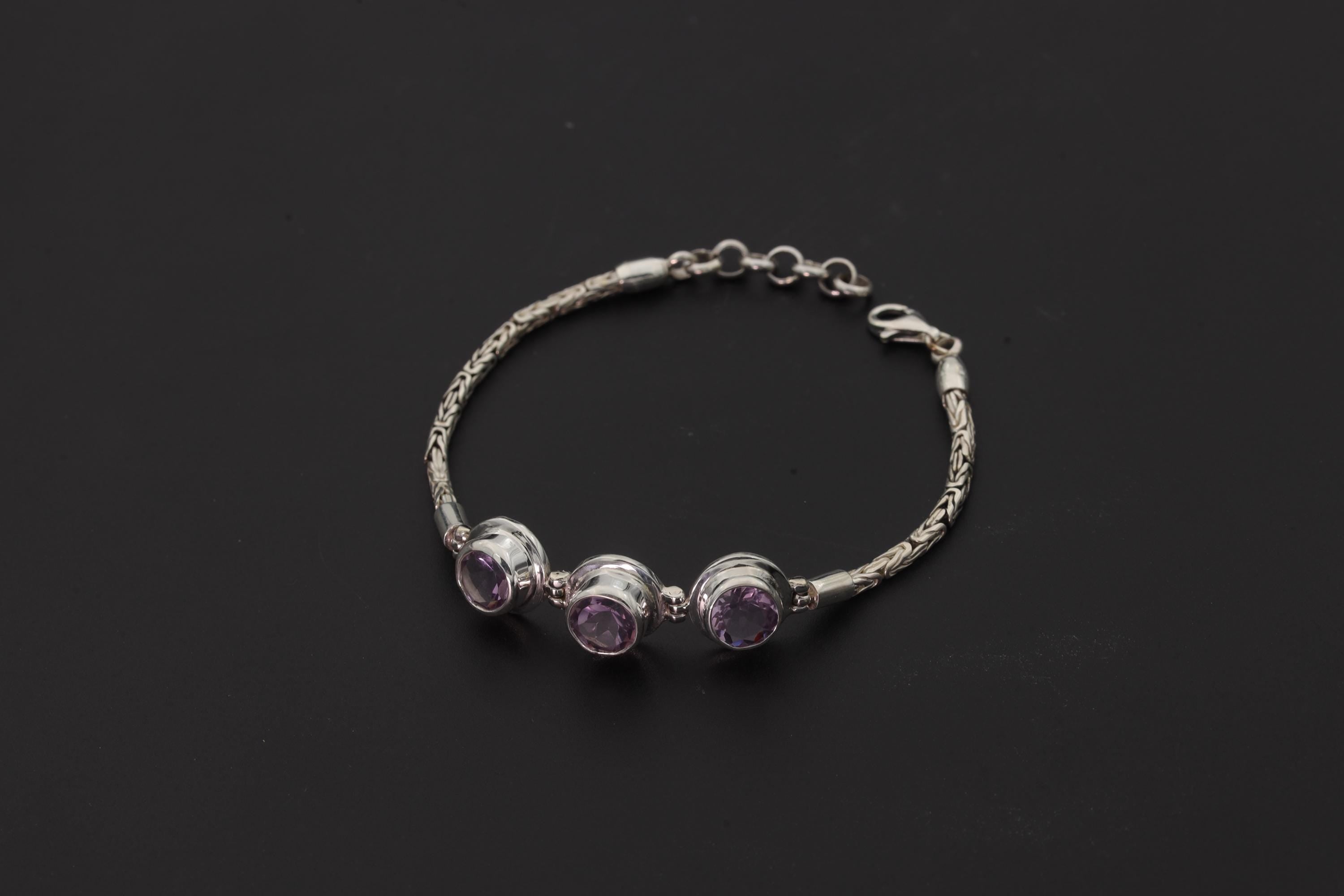 Amethyst bracelet displayed on dark background with three faceted purple stones and sterling silver Byzantine chain.