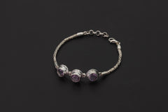 Amethyst bracelet displayed on dark background with three faceted purple stones and sterling silver Byzantine chain.
