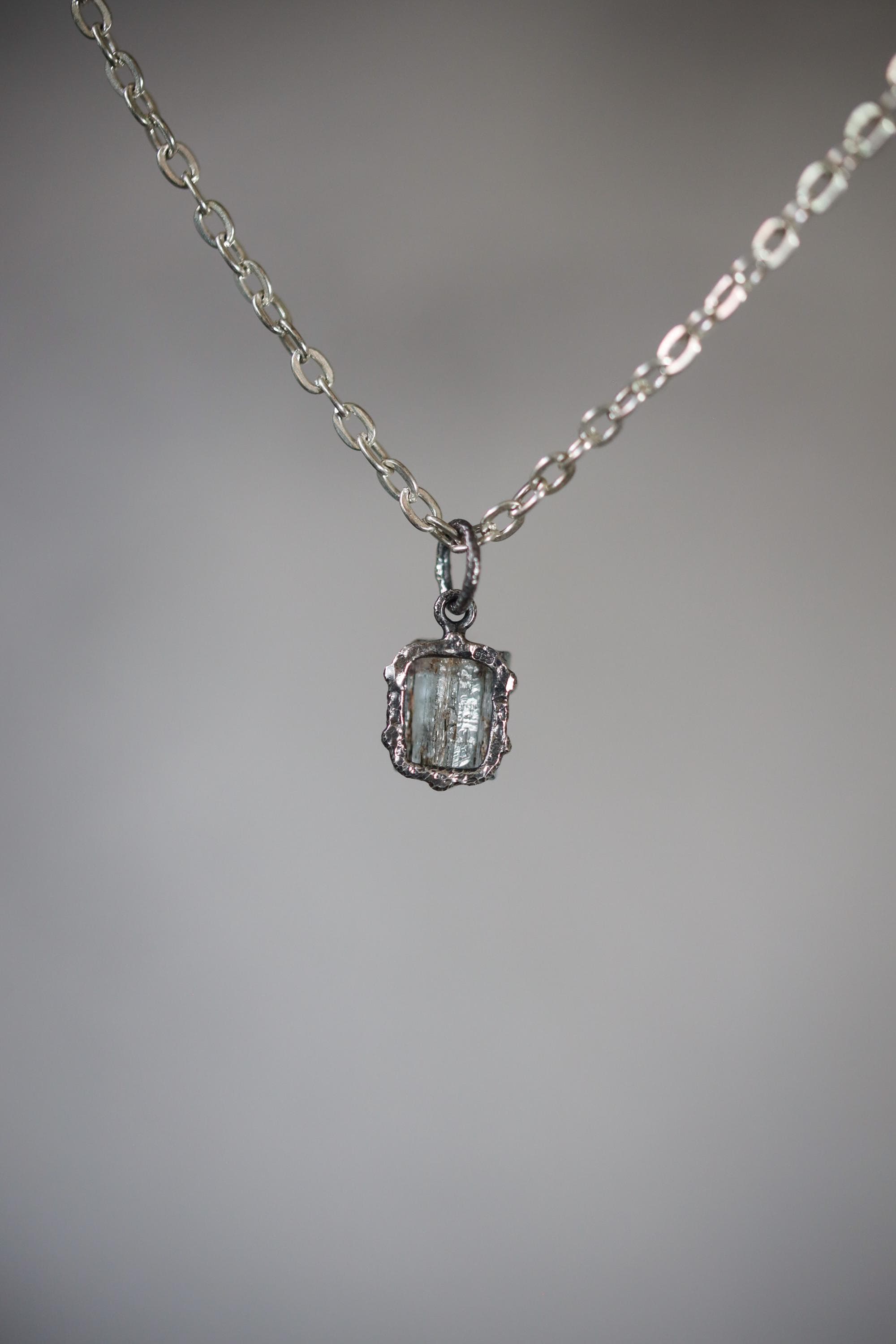 Aquamarine wand pendant shown on sterling silver chain with grey background, featuring raw blue-green crystal and claw setting.