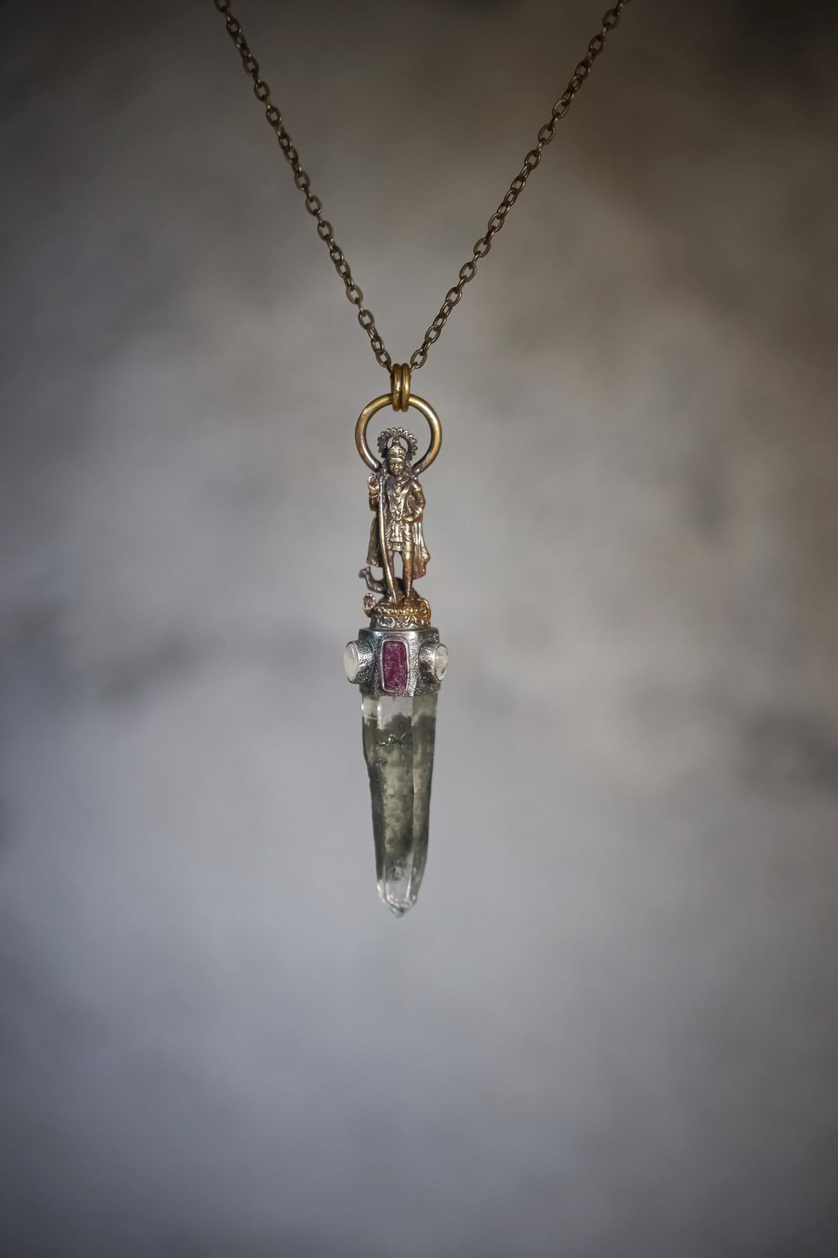 Saraswati Talisman Brass Cast Pendant on neutral gradient background showing brass Saraswati figure above Himalayan Chlorite Lemurian Quartz point with Rubellite and Blue gemstones in oxidized silver bezel.