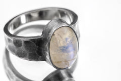 Close-up of AAA Blue Moonstone feldspar ring showing oval cabochon, oxidized silver bezel, and rugged hammered band.