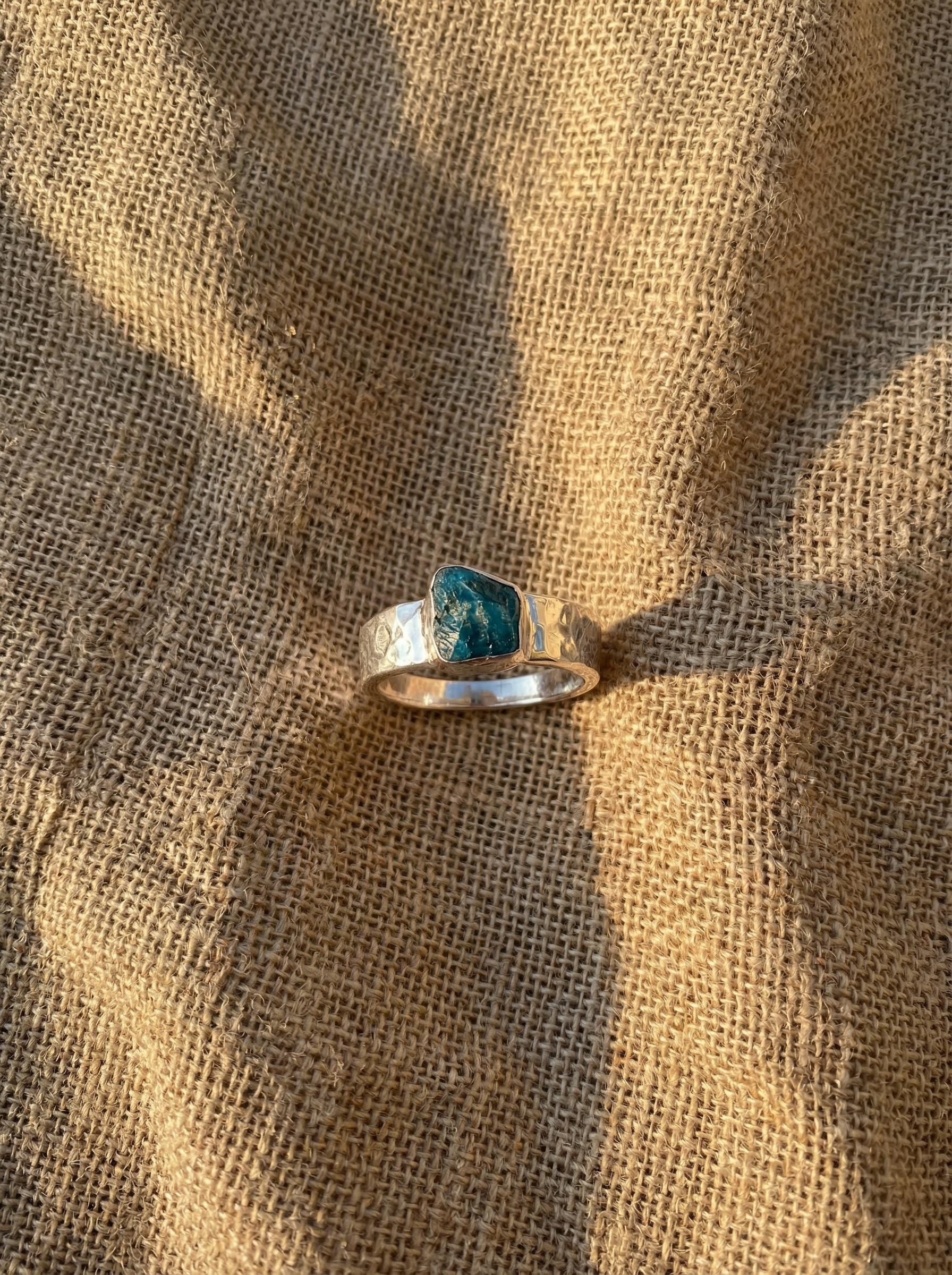 Blue Apatite Textured Silver Ring shown on sand-coloured fabric with bright blue gemstone and slim silver band.