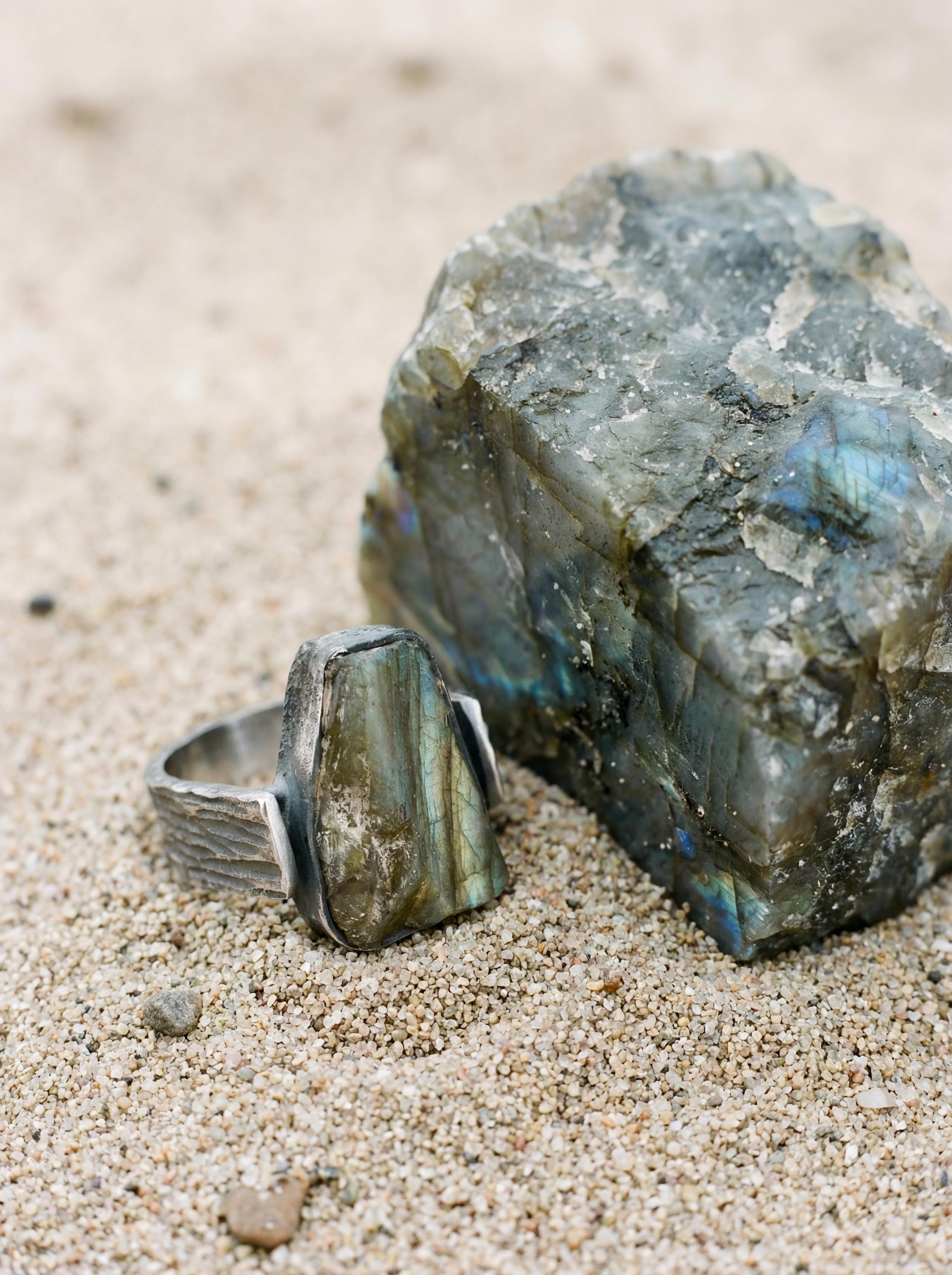Labradorite sterling silver ring shown on sand beside raw stone with blue-green mineral flash.
