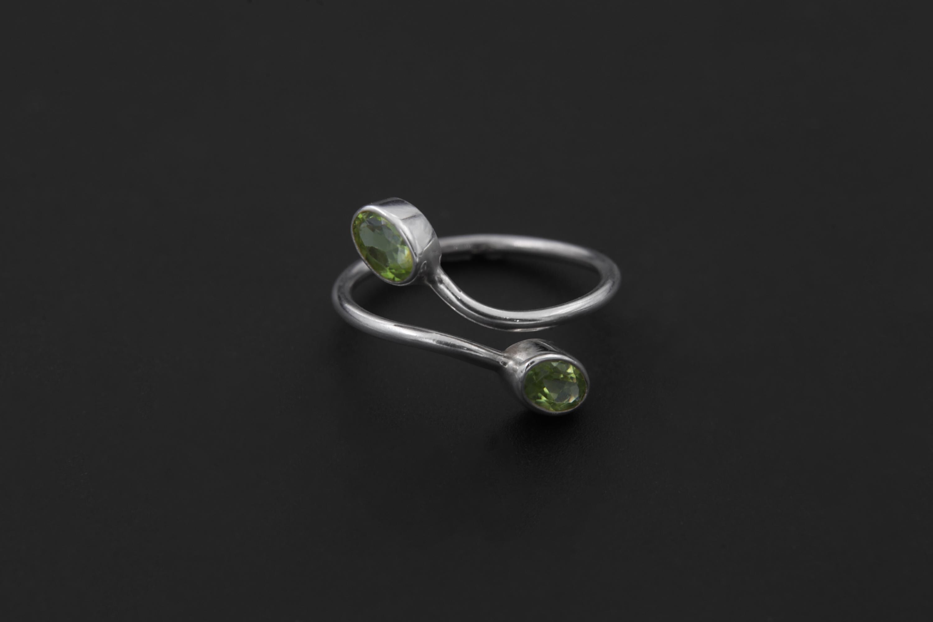 Peridot open wrap ring displayed on light stone with two green gemstones and sculptural silver band.
