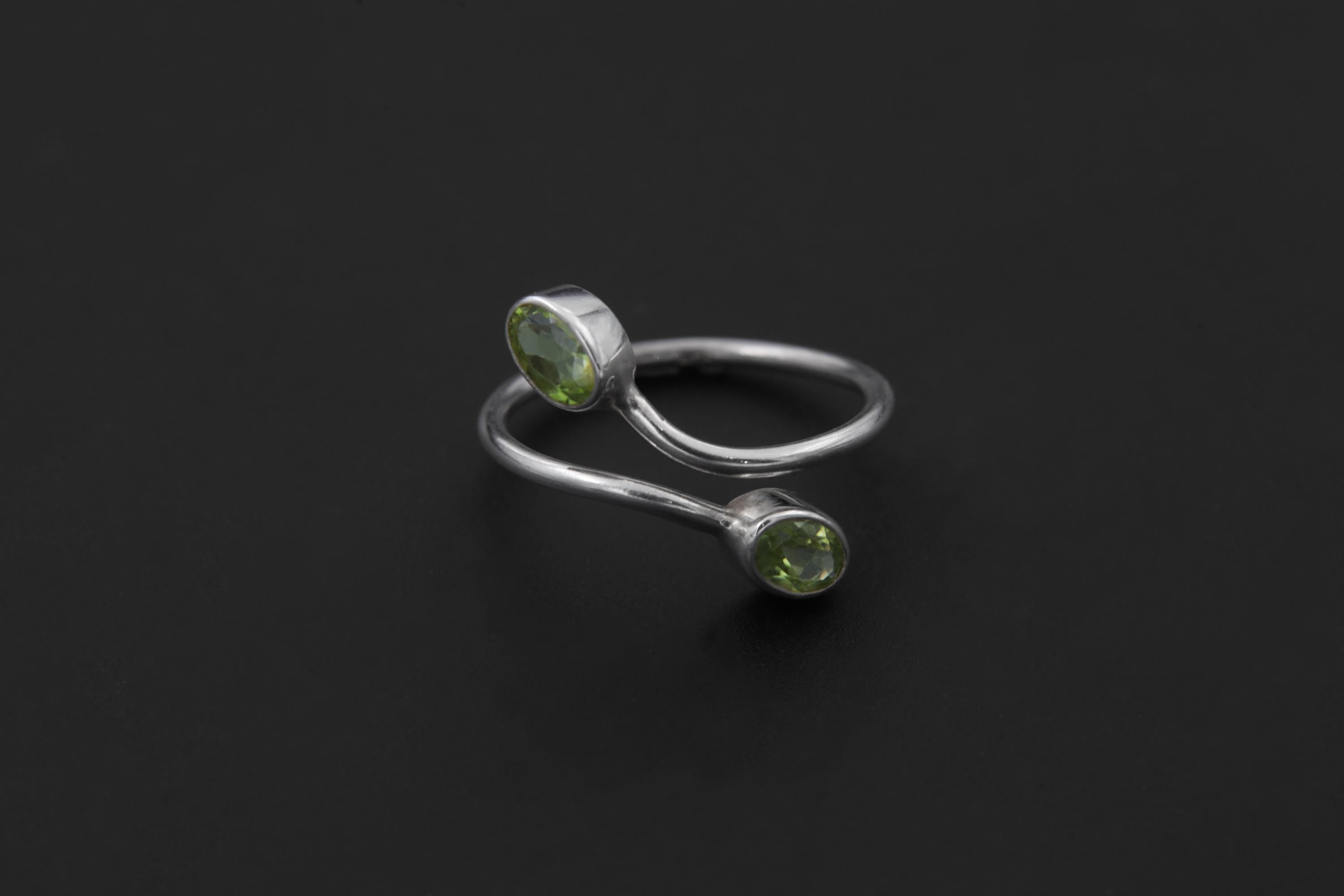Peridot open wrap ring displayed on light stone with two green gemstones and sculptural silver band.