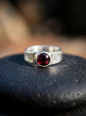 Crimson Sentinel Garnet Ring shown on dark surface with polished red Garnet and hammered sterling silver setting.