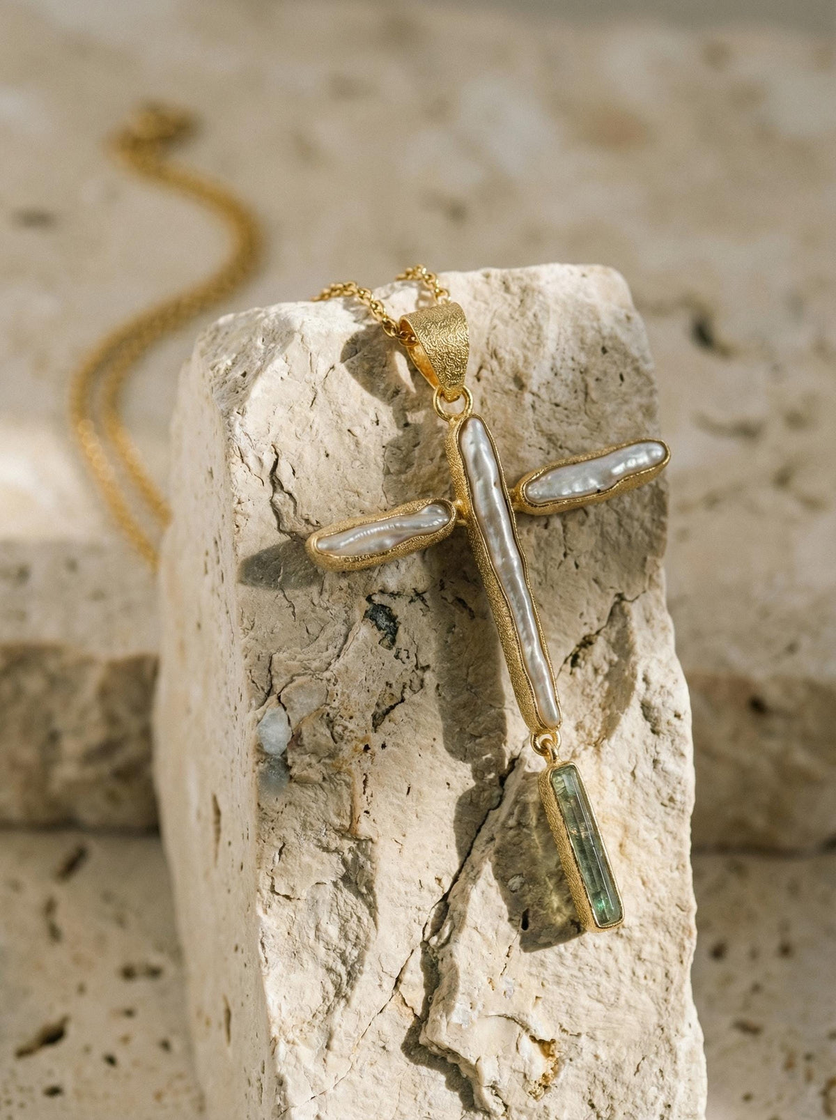 May include: A gold-colored cross pendant necklace with a textured finish. The cross features white, oblong pearls and a green rectangular gemstone at the bottom. The necklace is displayed on a light-colored, textured stone surface.